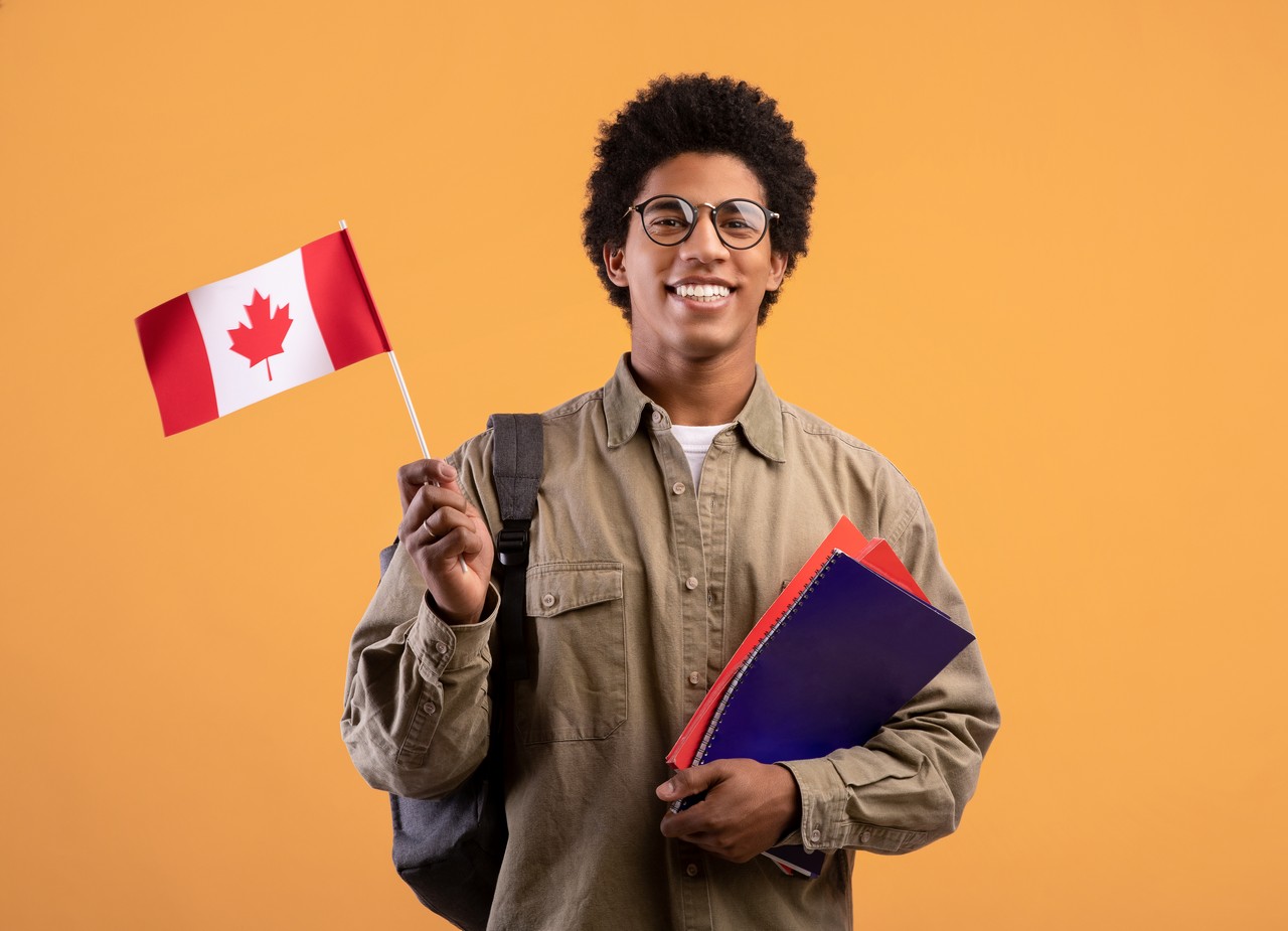 Education, school, college and university. Happy young african american guy in glasses holding notepads and small Canada flag and ready to study, isolated on orange background, free space, studio shot